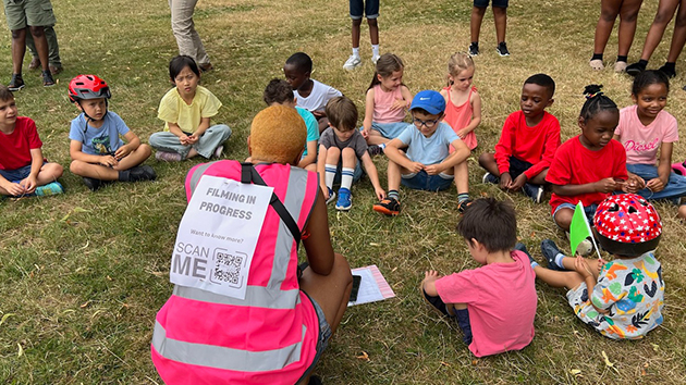 A Kingdom Kidz crew member speaks to the children during the music video shoot at Rothamsted Park, Harpenden. Her high-visibility vest features a QR code that passers-by can scan to learn more about the song and its message of hope.